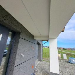 Damian Siwek - Nowoczesna elewacja domu z szarą, ziarnistą fakturą i białym podbitką. Widoczne detale wykończenia, przewody i niebieski wąż. Tło stanowi zielony krajobraz.