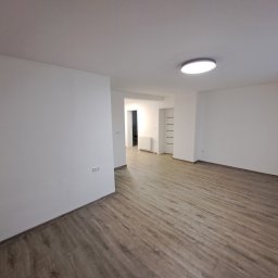 Damian Siwek - Jasne, puste wnętrze po remoncie: parkiet w odcieniach brązu, białe ściany i sufit z okrągłą lampą. Widok na korytarz z grzejnikiem i białymi drzwiami.