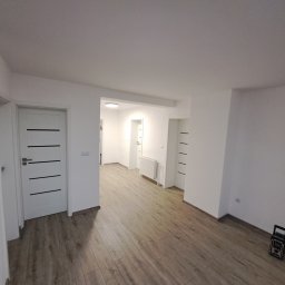 Damian Siwek - Jasne, przestronne wnętrze po remoncie: świeżo ułożone panele podłogowe, białe ściany i drzwi z poziomymi, czarnymi akcentami. Widoczny grzejnik i przenośne radio.