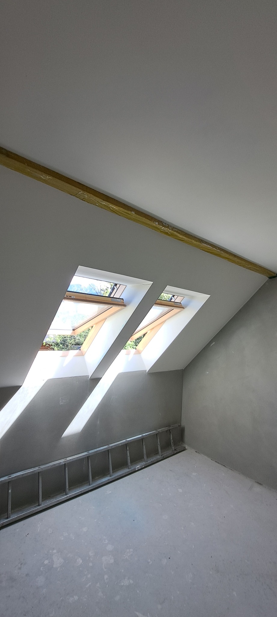 Poddasze po remoncie z dwoma oknami dachowymi, przez które wpada światło, widoczna drabina oparta o ścianę, ściany przygotowane do malowania, belka zabezpieczona folią.