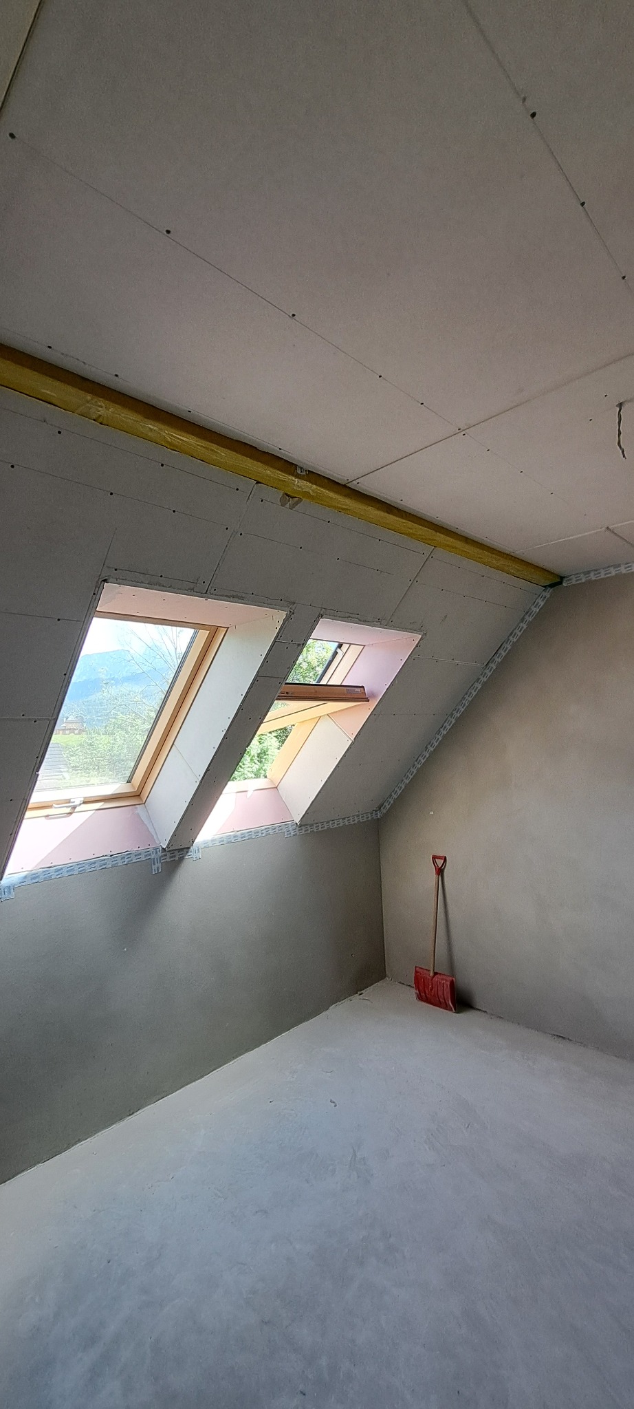 Poddasze z oknami dachowymi w trakcie wykańczania, widoczne płyty gipsowo-kartonowe, ocieplenie wełną mineralną i czerwona łopata oparta o ścianę.