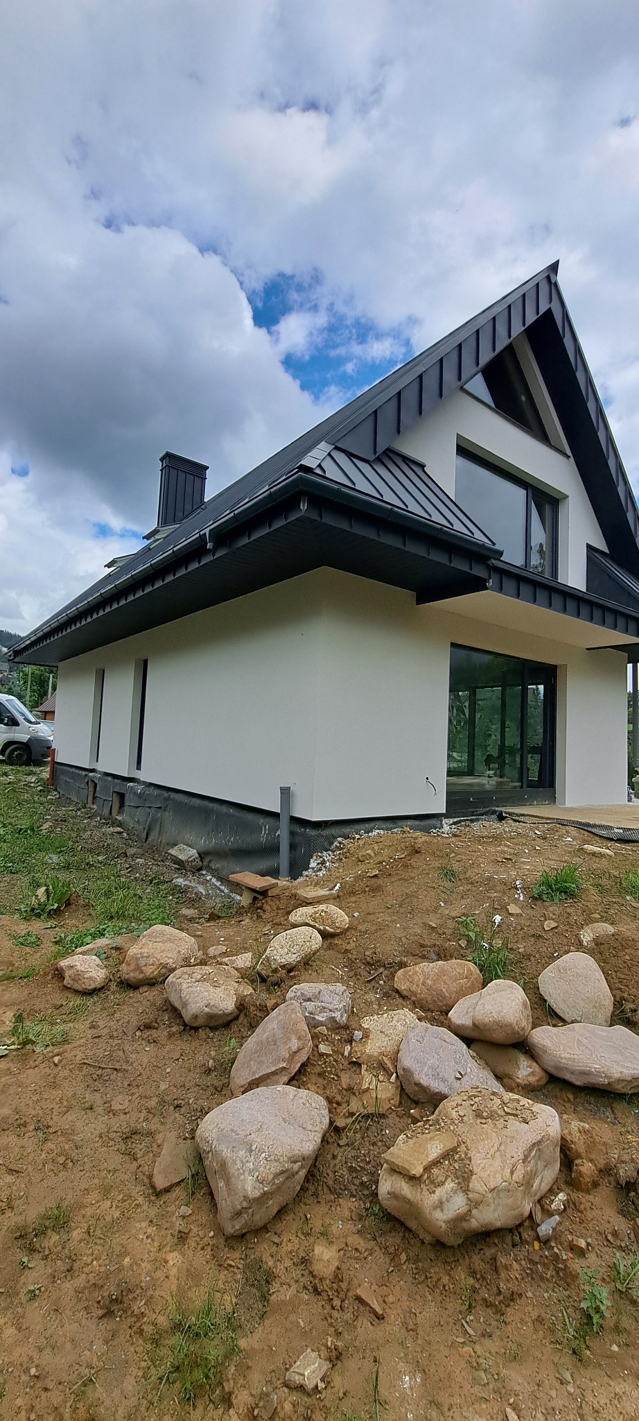 Nowoczesny dom z białą elewacją i czarnym dachem z blachy na tle pochmurnego nieba, widoczne kamienie w ziemi przed budynkiem.