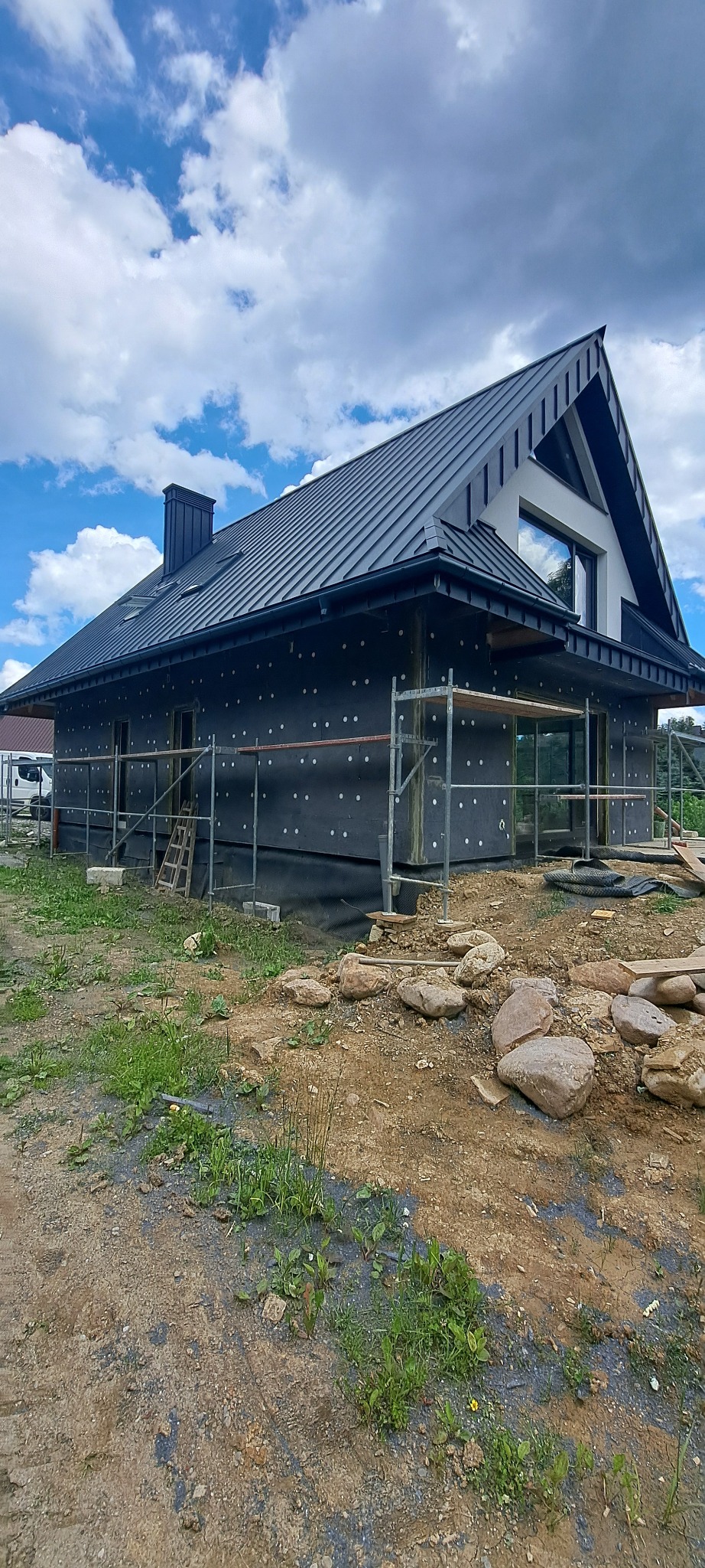 Dom jednorodzinny w trakcie ocieplania, widoczne rusztowania, czarna membrana z białymi kropkami na elewacji, ciemny dach z blachy na tle błękitnego nieba z chmurami.