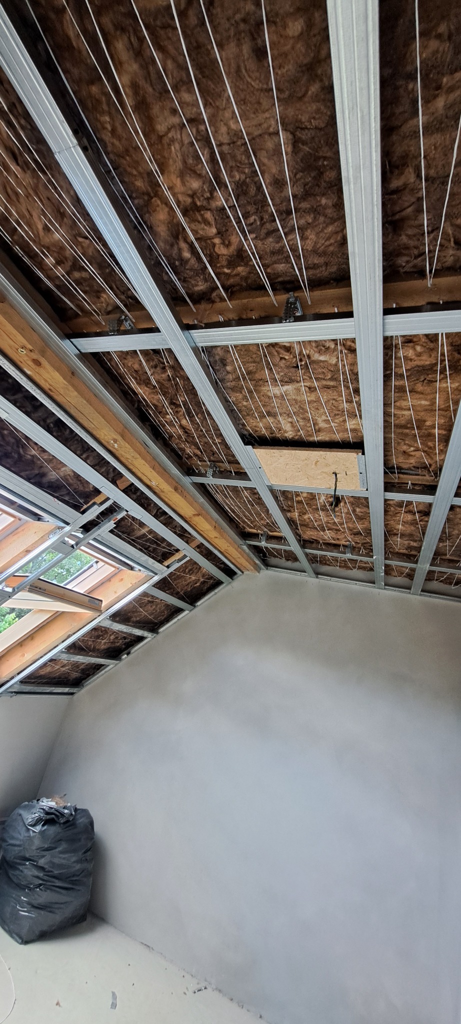 Poddasze w trakcie ocieplania: widoczne wełna mineralna, metalowy stelaż, sznurki podtrzymujące izolację oraz okno dachowe, ściany częściowo wykończone gładzią, na podłodze czarny worek na śmieci.