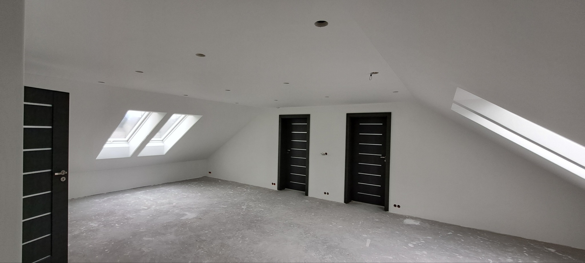 Poddasze po remoncie z dwiema ciemnymi, nowoczesnymi drzwiami, oknami dachowymi i przygotowaną podłogą, widoczne ślady po instalacjach elektrycznych.