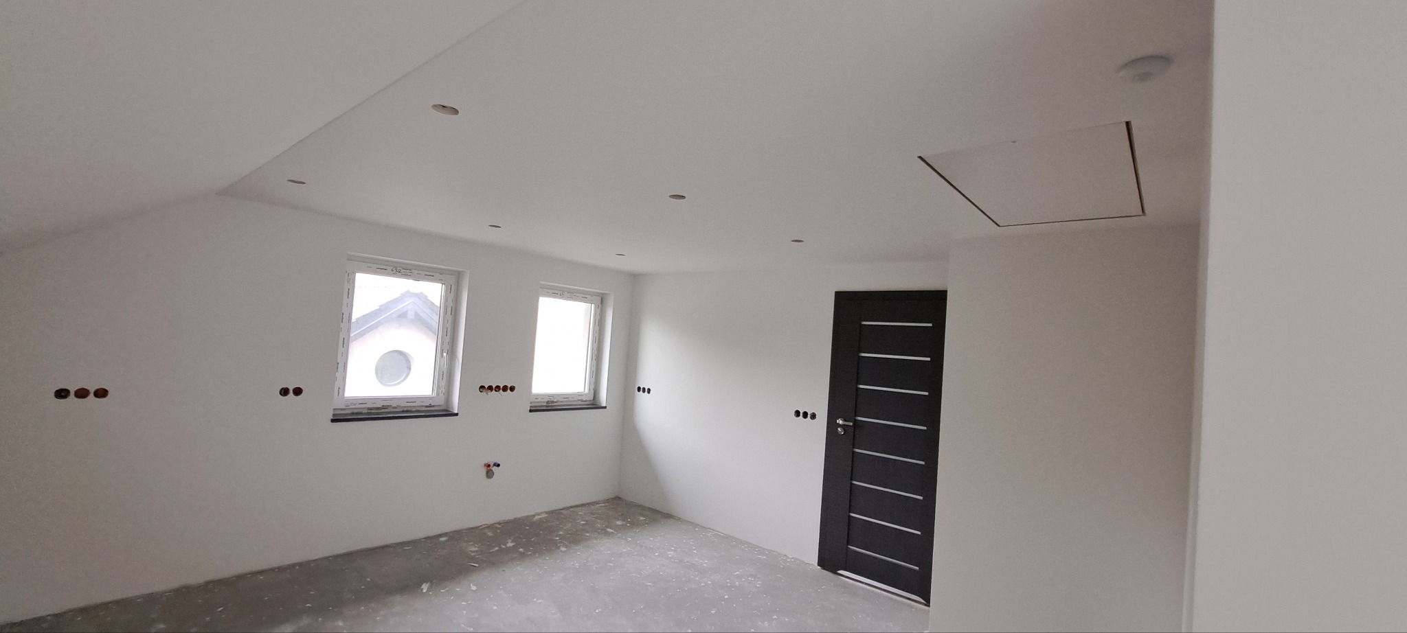 Wnętrze pokoju na poddaszu po remoncie, z dwiema oknami, czarnymi drzwiami z poziomymi paskami, przygotowanymi punktami elektrycznymi na ścianach i surową podłogą przed położeniem paneli.