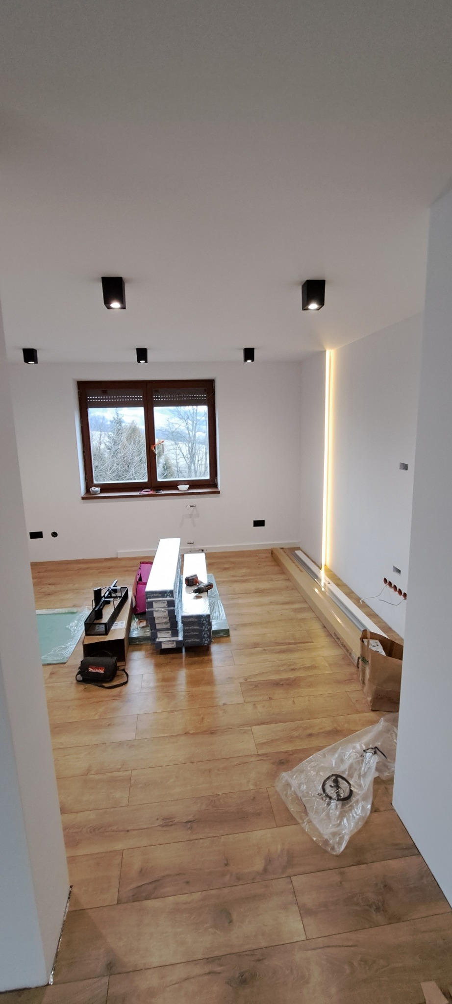 Pokój w trakcie układania paneli podłogowych z widocznymi paczkami paneli, narzędziami i oświetleniem LED na ścianie.