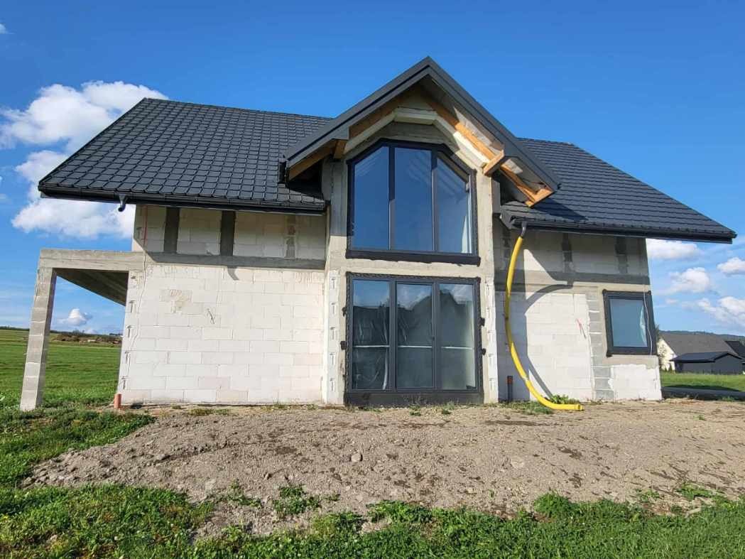 Dom w trakcie budowy z szarym dachem i dużymi oknami. Ściany z białych bloczków, widoczne elementy konstrukcyjne. Żółty wąż przy ścianie. Błękitne niebo w tle.