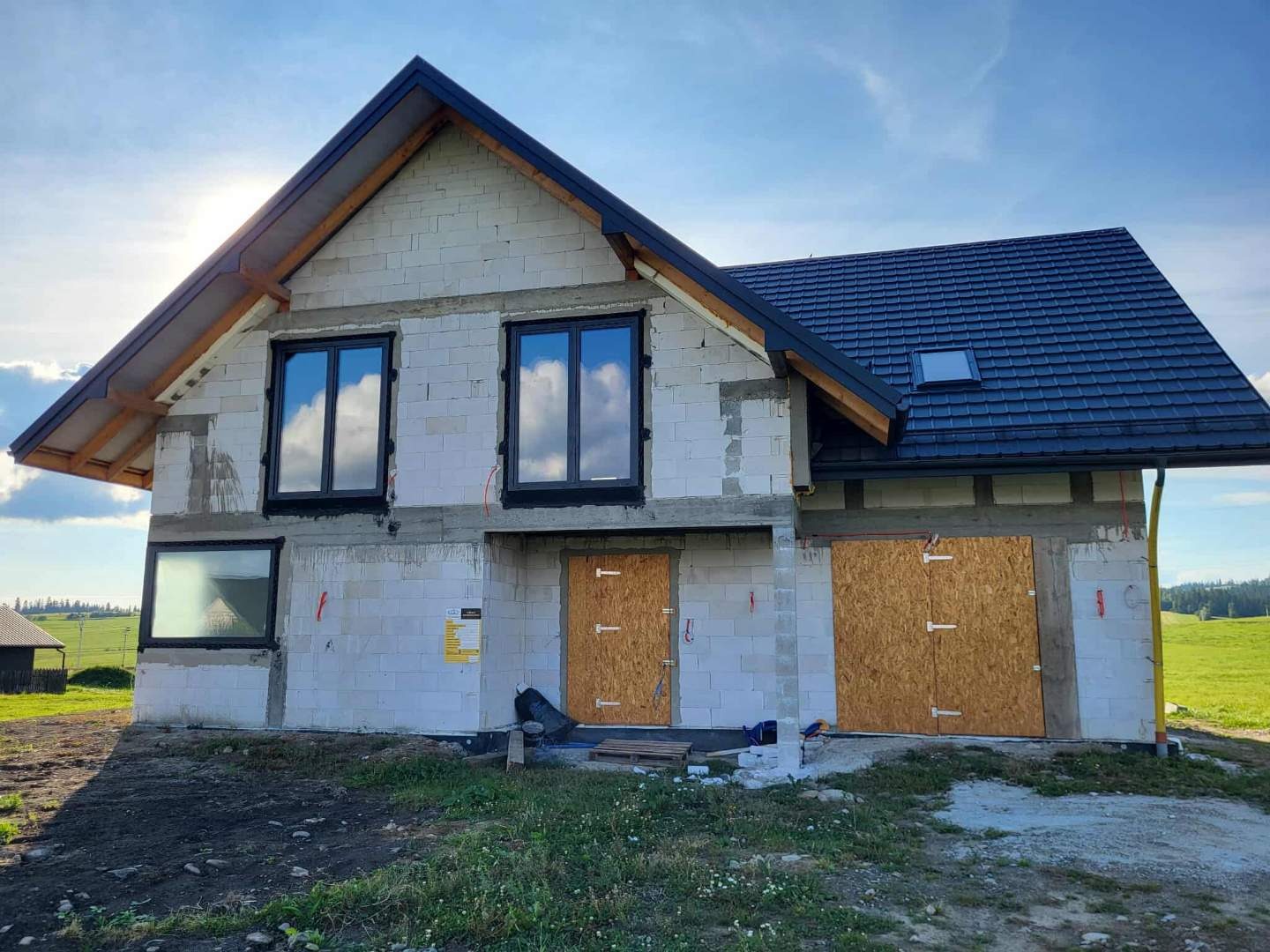 Niedokończony dom jednorodzinny z czarną dachówką i oknami, ściany z białych bloczków, zabezpieczone wejścia, błękitne niebo z chmurami odbija się w szybach.