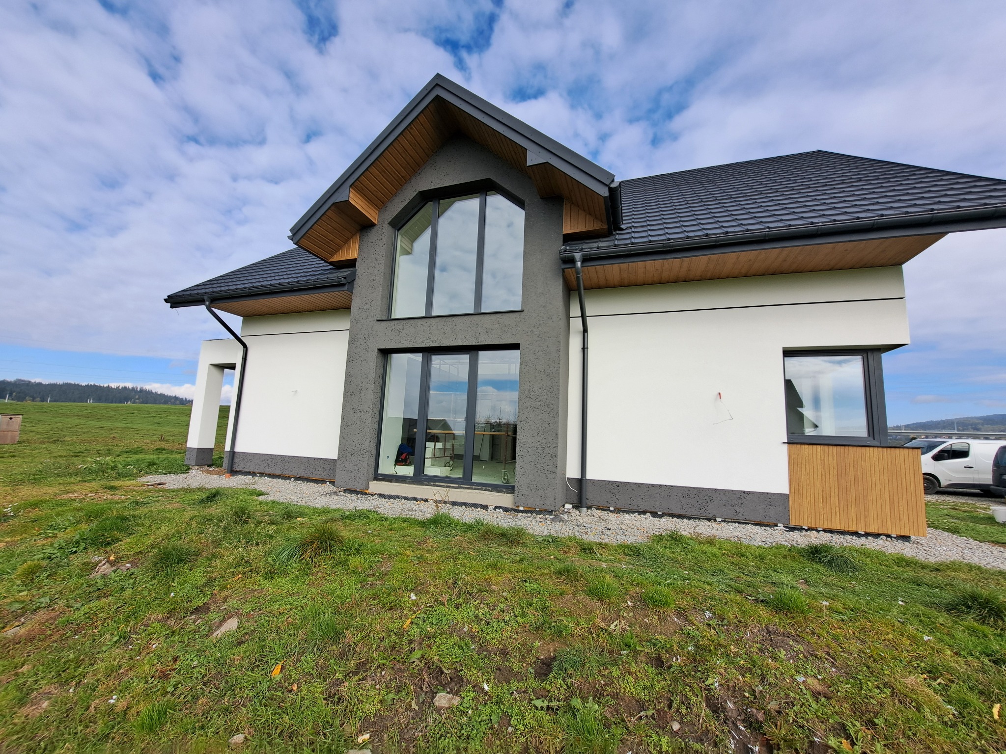 Nowoczesny dom jednorodzinny z dużymi oknami i elewacją w odcieniach bieli i szarości, otoczony zielonym trawnikiem i błękitnym niebem z chmurami.