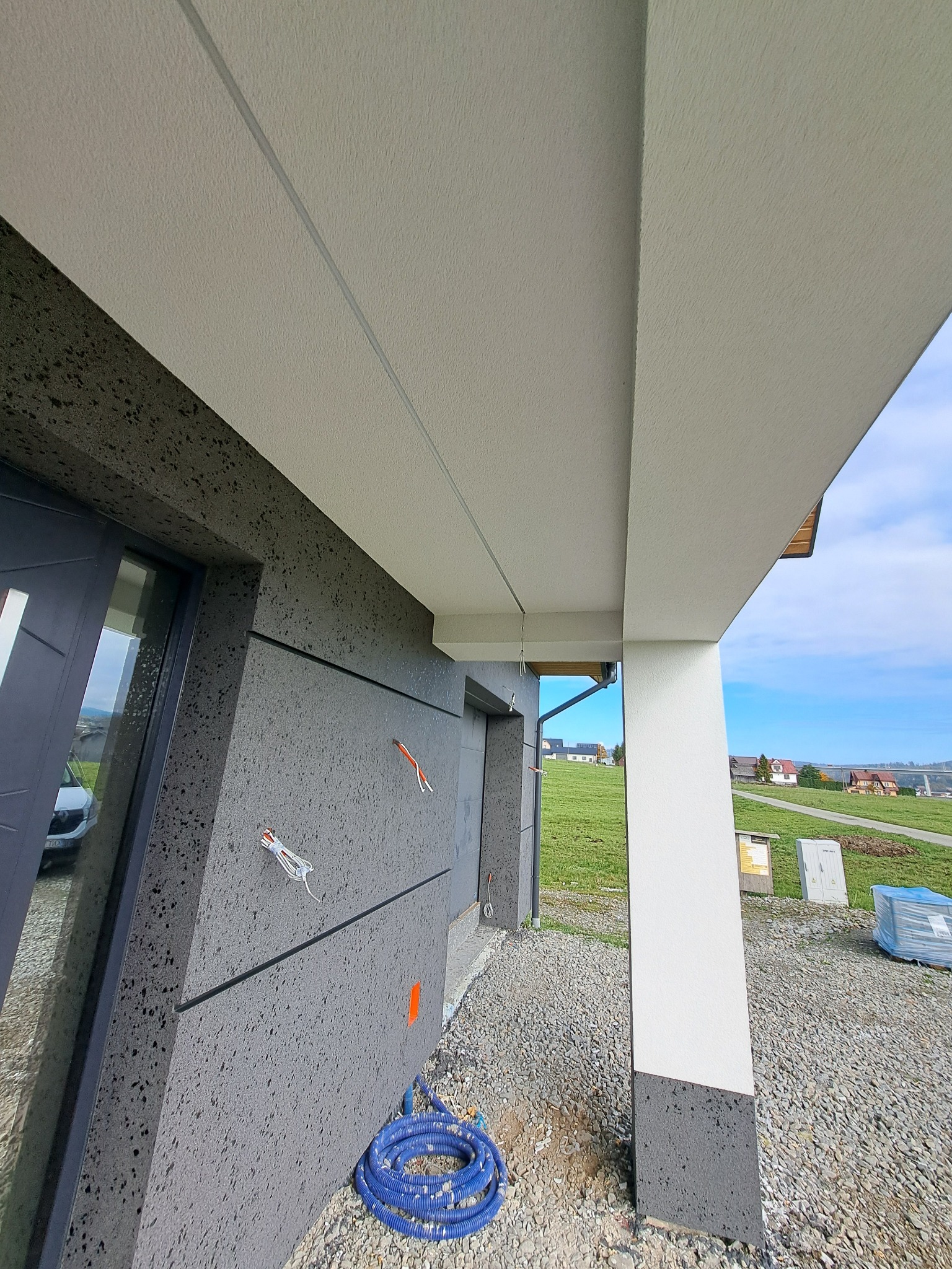 Nowoczesna elewacja domu z szarą, ziarnistą fakturą i białym podbitką. Widoczne detale wykończenia, przewody i niebieski wąż. Tło stanowi zielony krajobraz.