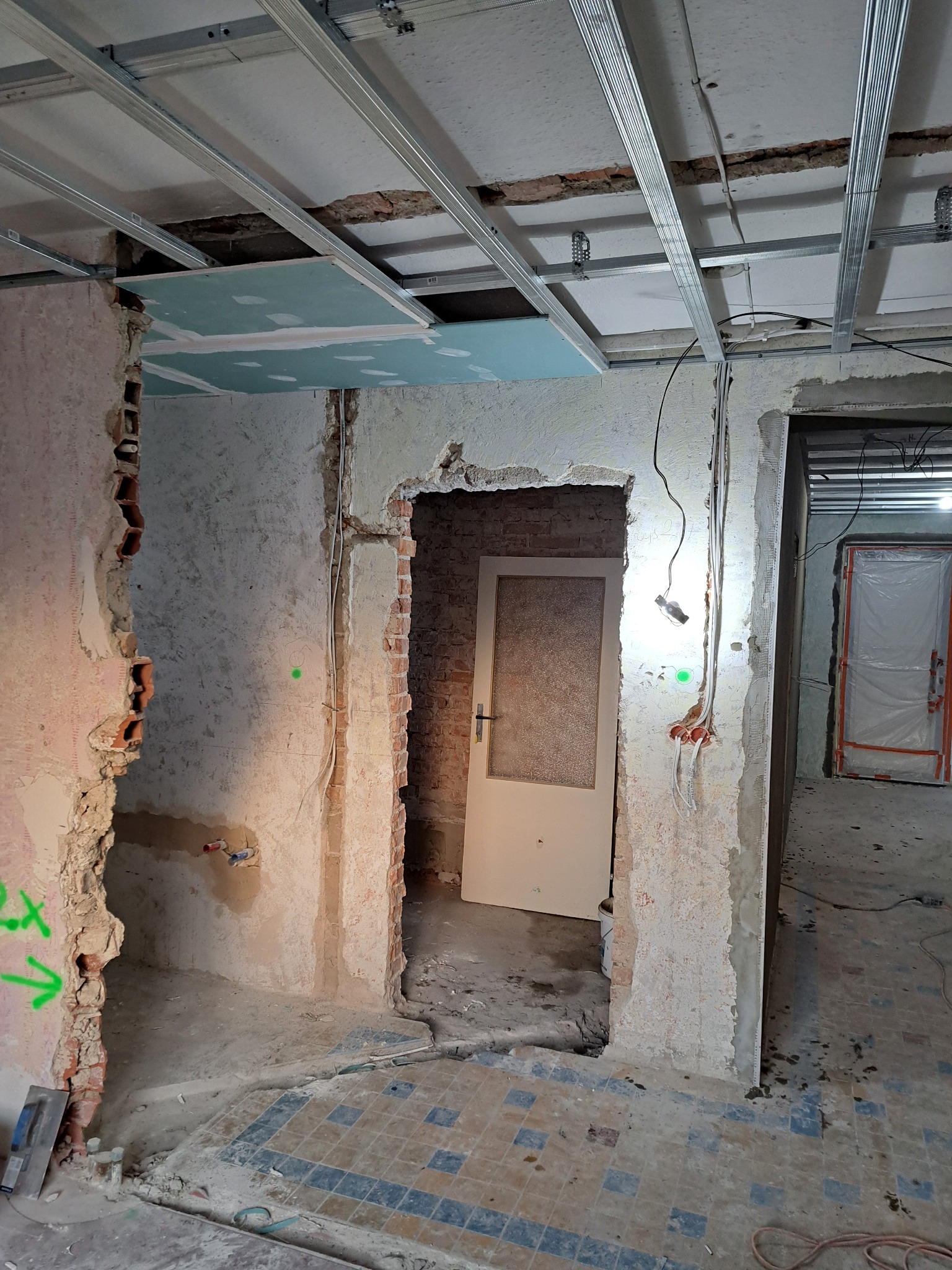 Wnętrze w trakcie prac: widoczne metalowe profile sufitowe, fragment płyty, odsłonięta cegła, instalacje elektryczne i fragment podłogi z płytek. Przejście do kolejnego pomieszczenia.