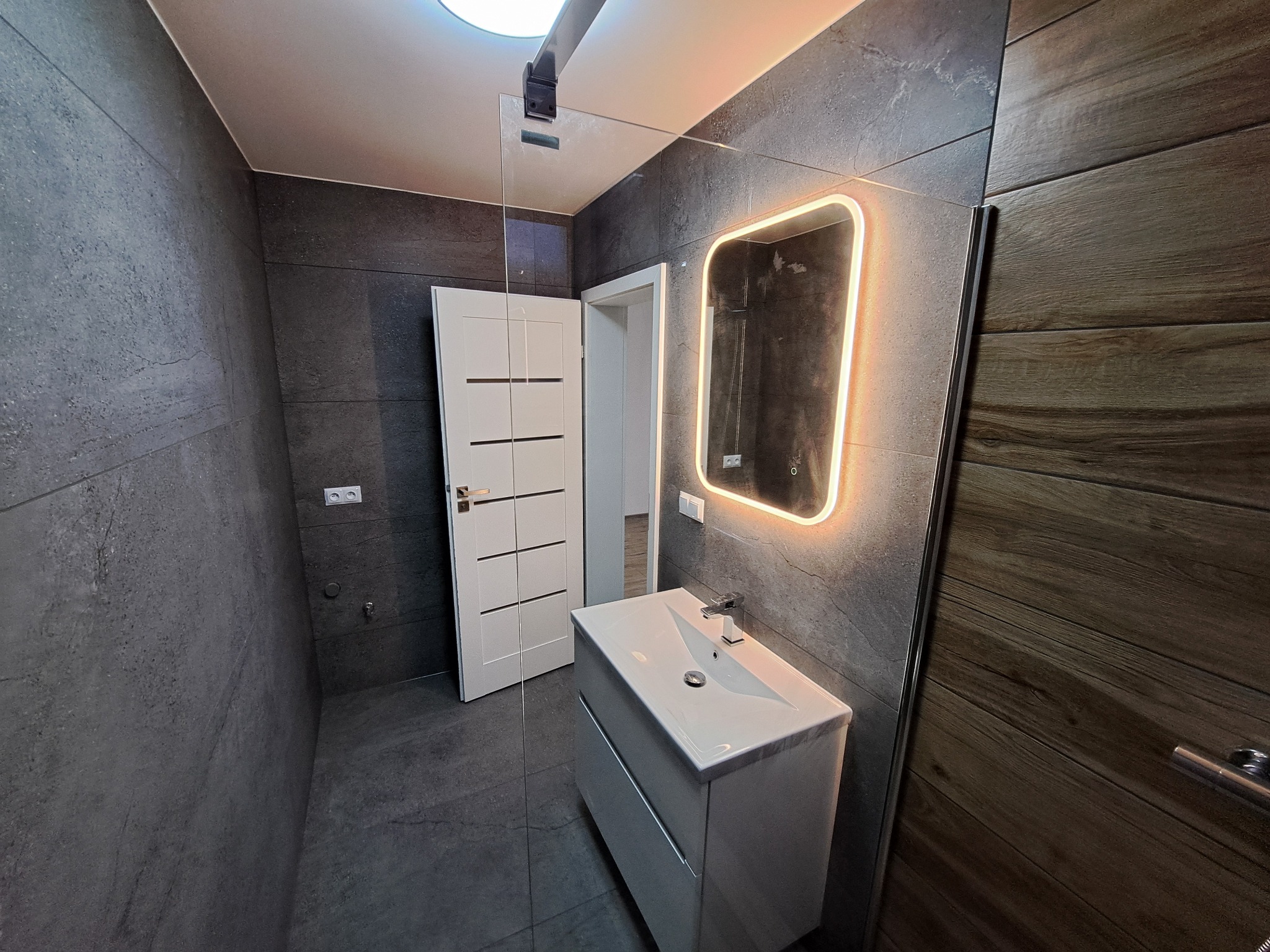 Nowoczesna łazienka z szarymi płytkami imitującymi kamień, podświetlanym lustrem i białą szafką z umywalką. Widoczna biała, wewnętrzna stolarka drzwiowa.