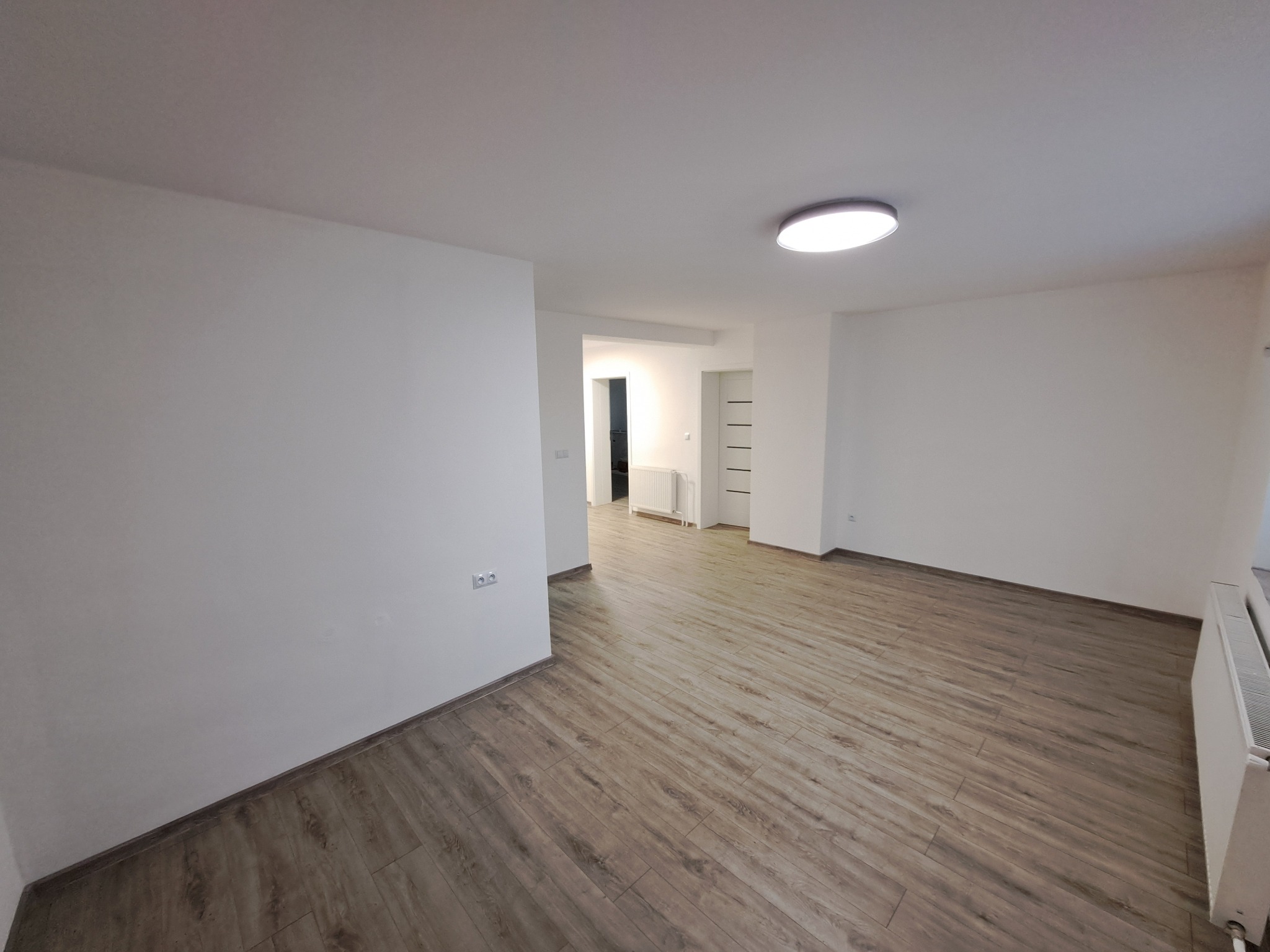 Jasne, puste wnętrze po remoncie: parkiet w odcieniach brązu, białe ściany i sufit z okrągłą lampą. Widok na korytarz z grzejnikiem i białymi drzwiami.