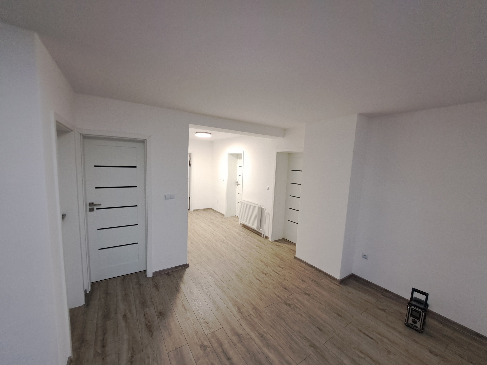 Jasne, przestronne wnętrze po remoncie: świeżo ułożone panele podłogowe, białe ściany i drzwi z poziomymi, czarnymi akcentami. Widoczny grzejnik i przenośne radio.