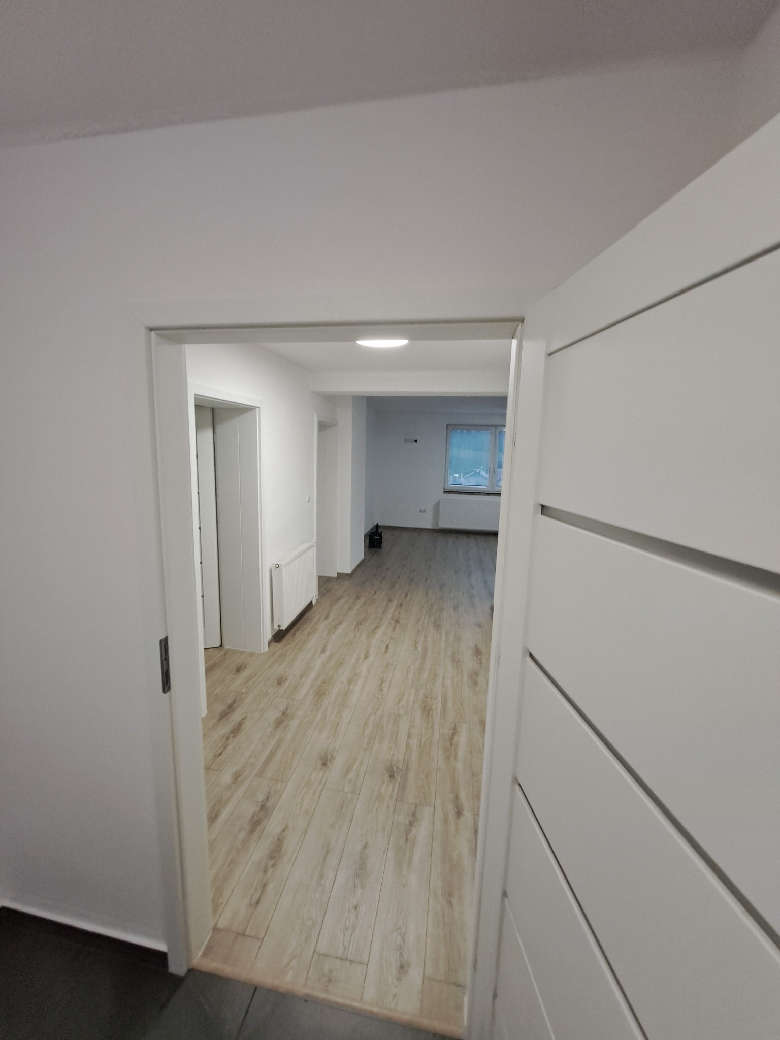 Jasne wnętrze po remoncie: panele podłogowe, białe ściany i drzwi z poziomymi frezami. Widok z korytarza do pokoju z oknem. Minimalistyczny design.