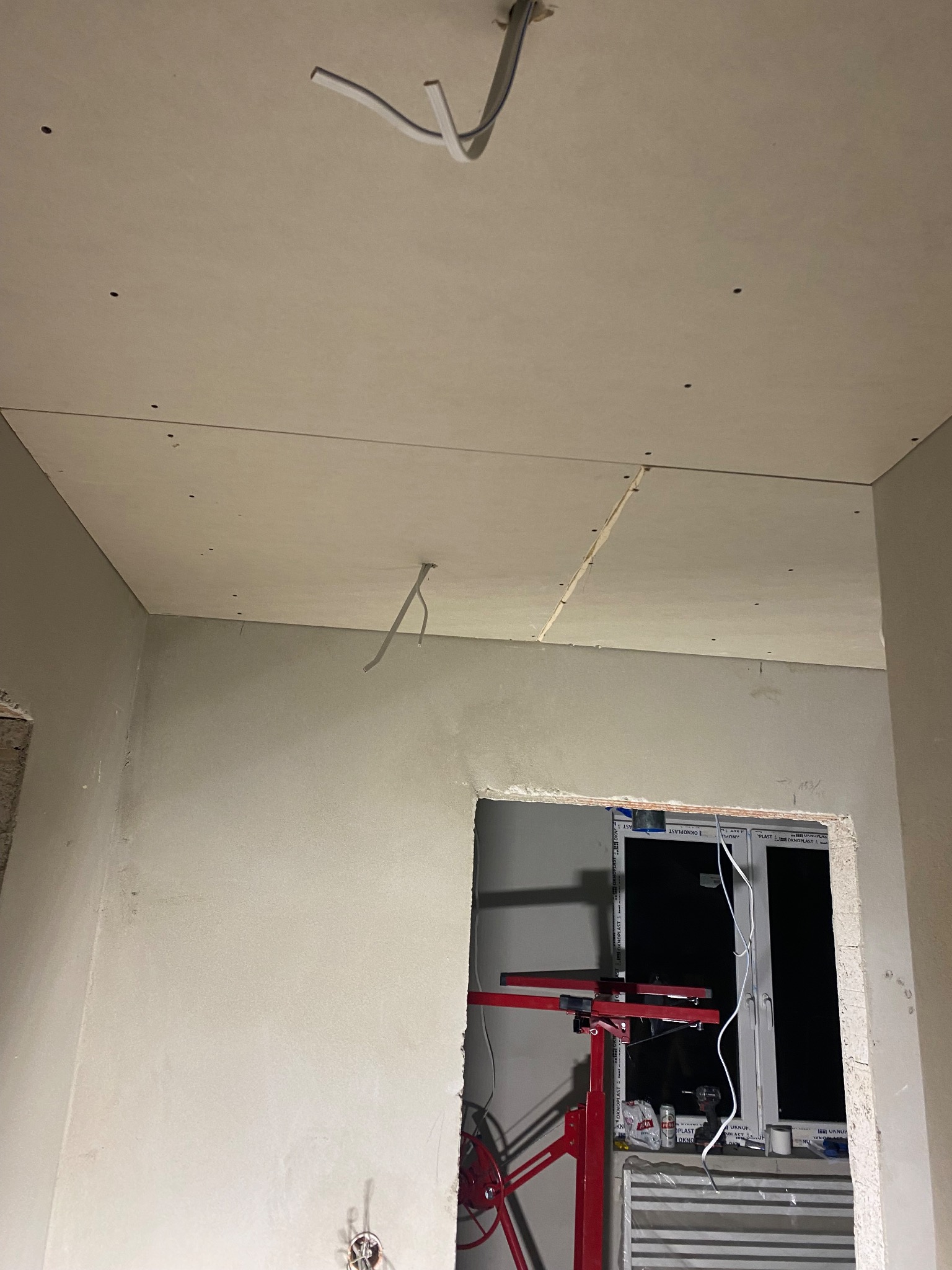 Montaż płyty gipsowo-kartonowej na suficie, widoczne kable elektryczne i podnośnik do płyt, surowe ściany przygotowane do malowania.