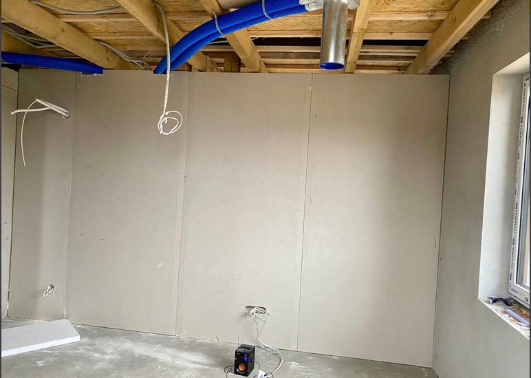 Wnętrze w trakcie wykańczania: ściany z płyt kartonowo-gipsowych, widoczne przewody wentylacyjne pod sufitem, instalacja elektryczna wyprowadzona ze ściany, surowy beton na podłodze.