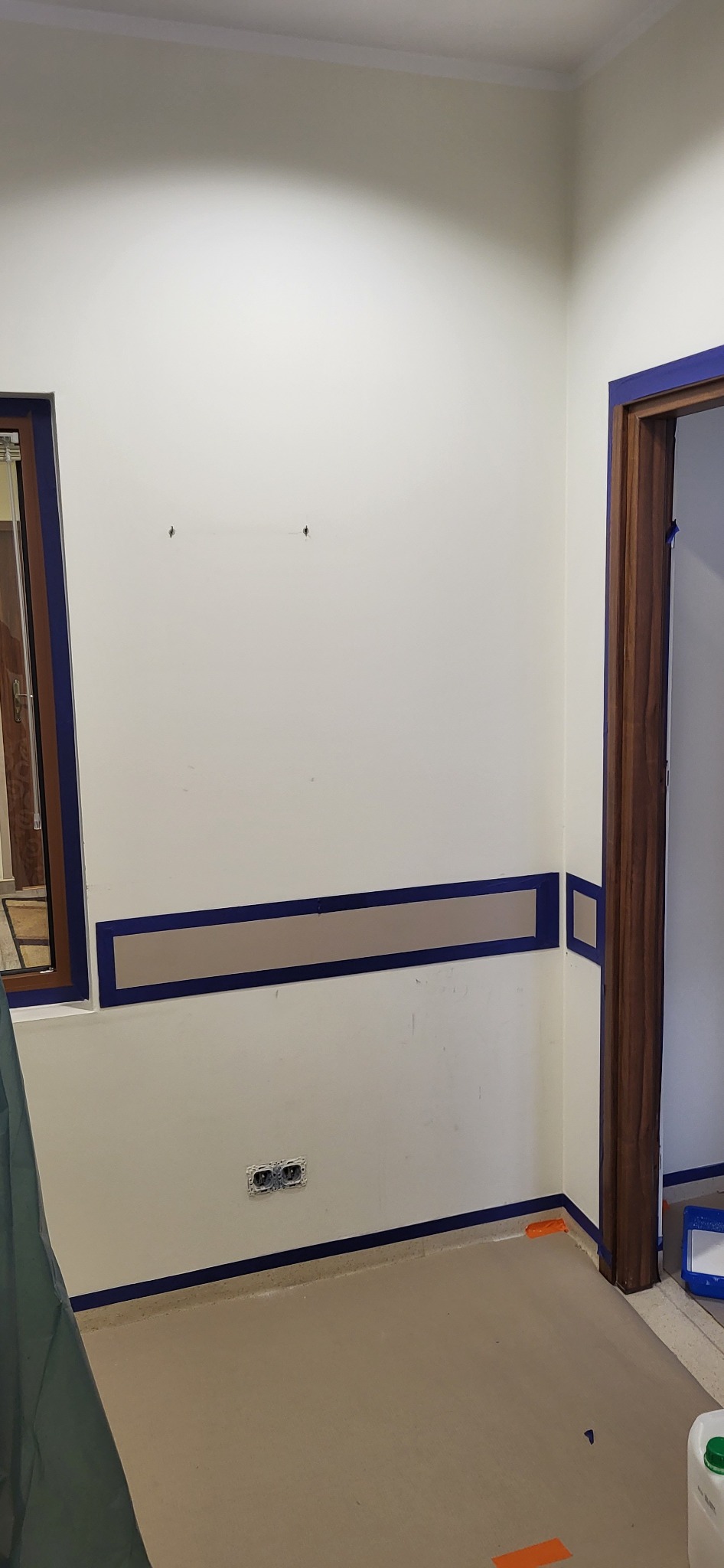 Przygotowania do malowania pokoju: ściany zabezpieczone niebieską taśmą malarską, listwa dekoracyjna pomalowana na brązowo, widoczne gniazdka elektryczne i fragment podłogi zabezpieczonej folią.