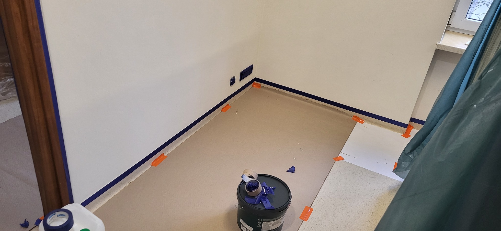 Przygotowane do malowania wnętrze pokoju z zabezpieczonymi listwami przypodłogowymi taśmą malarską, widoczny kubeł z farbą i rękawicami oraz folia ochronna na podłodze.