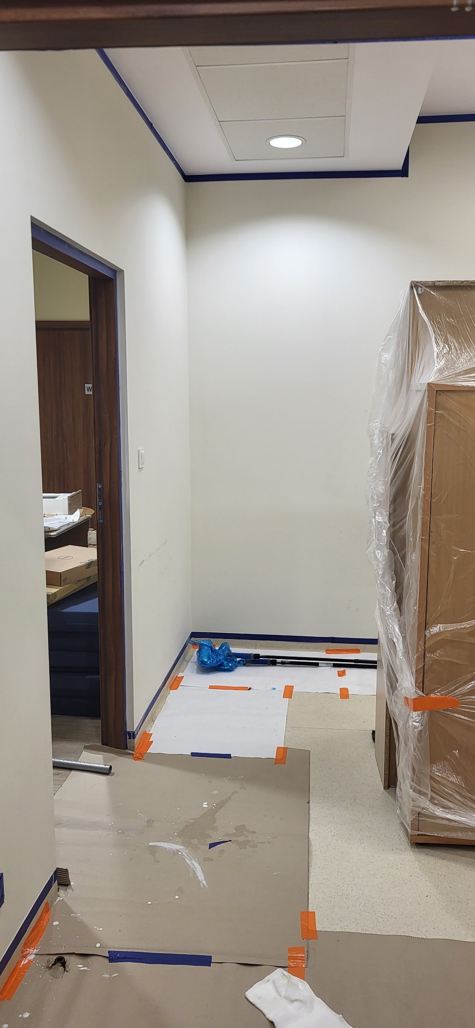 Przygotowane pomieszczenie do prac malarskich: zabezpieczone folią meble, taśmą malarską odcięte ściany i sufit, rozłożona tektura na podłodze z widocznymi śladami farby.