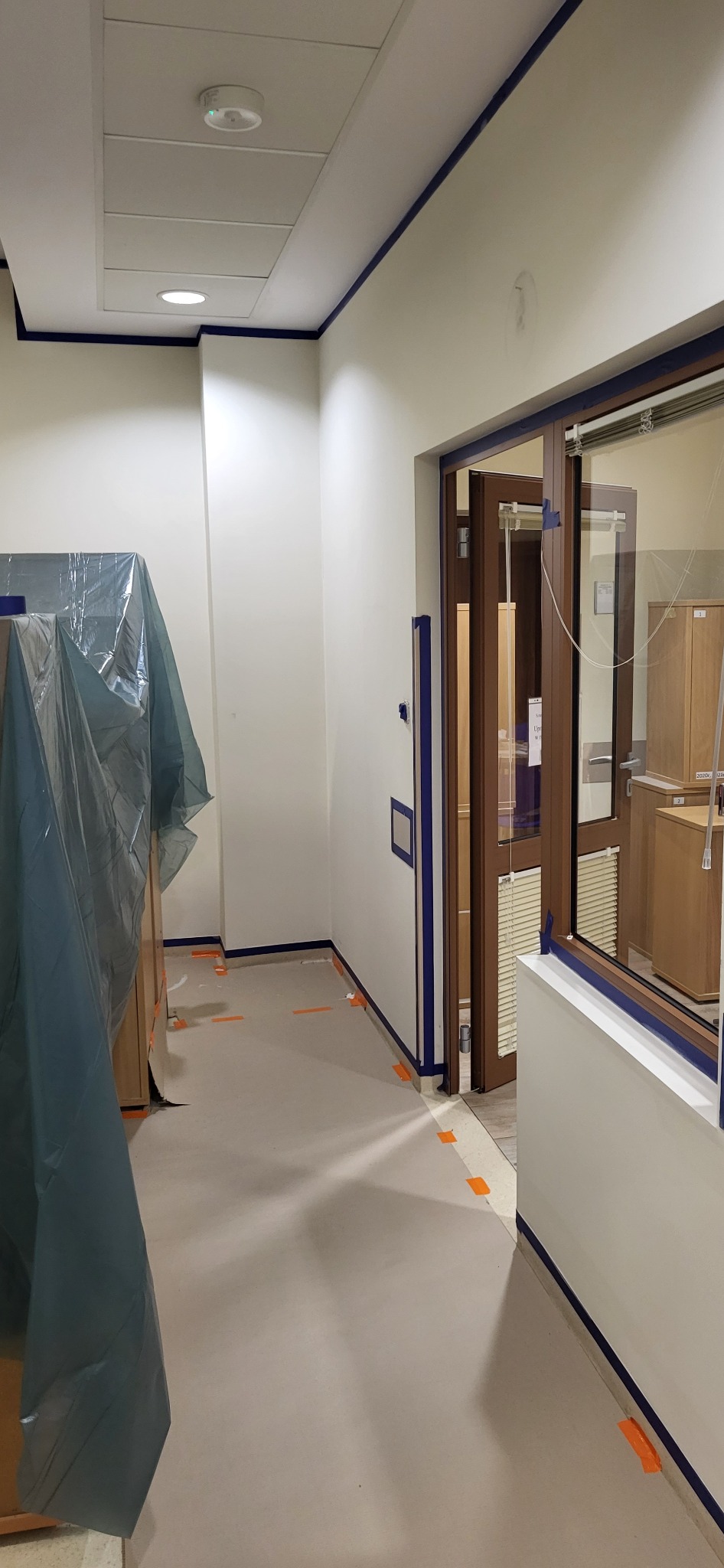 Przygotowanie do malowania w biurze: ściany i framugi oklejone taśmą malarską, meble zabezpieczone folią ochronną.