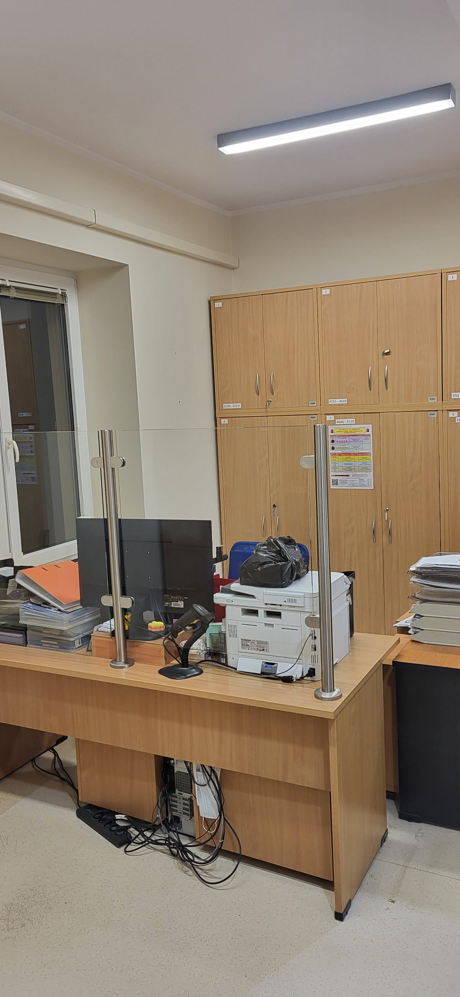 Biuro z zamontowaną szklaną przegrodą ochronną na biurku, monitor, drukarka, skaner kodów kreskowych i szafki w tle.