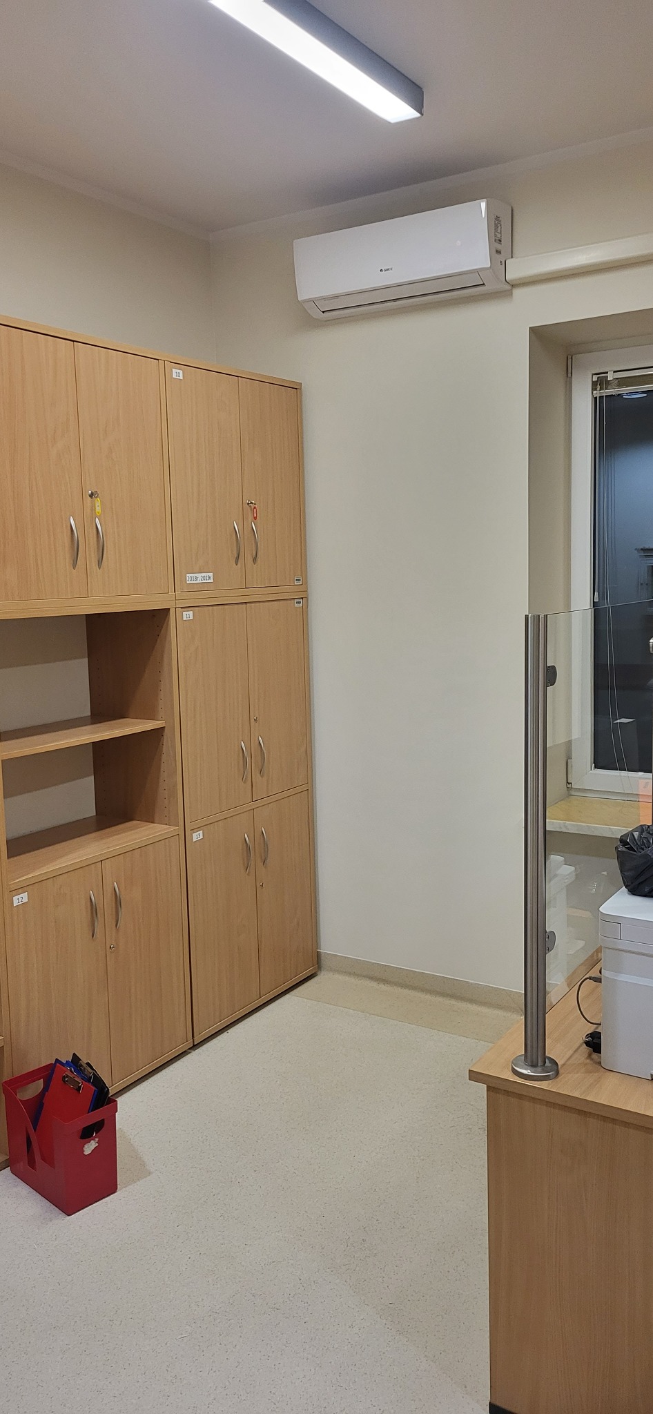 Wnętrze biura z jasnymi drewnianymi szafkami z numerami, klimatyzatorem na ścianie i czerwonym pojemnikiem z dokumentami na podłodze.