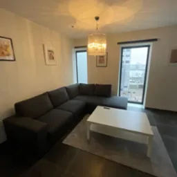 Szary narożnik z niskim stolikiem kawowym na tle okien z widokiem na miasto, oświetlony ozdobną lampą wiszącą.