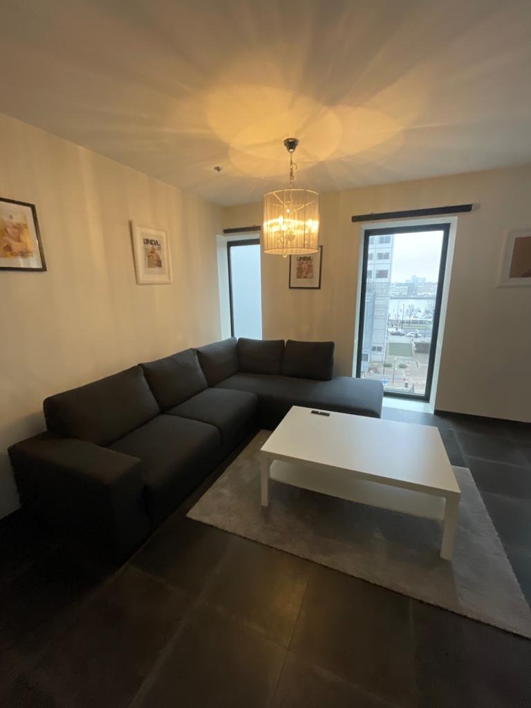 Szary narożnik z niskim stolikiem kawowym na tle okien z widokiem na miasto, oświetlony ozdobną lampą wiszącą.