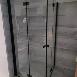 Tomasz Rychlik - Nowoczesna kabina prysznicowa z czarnymi profilami i przezroczystym szkłem, zamontowana w łazience z szarymi płytkami. Widoczny fragment drabiny w tle.