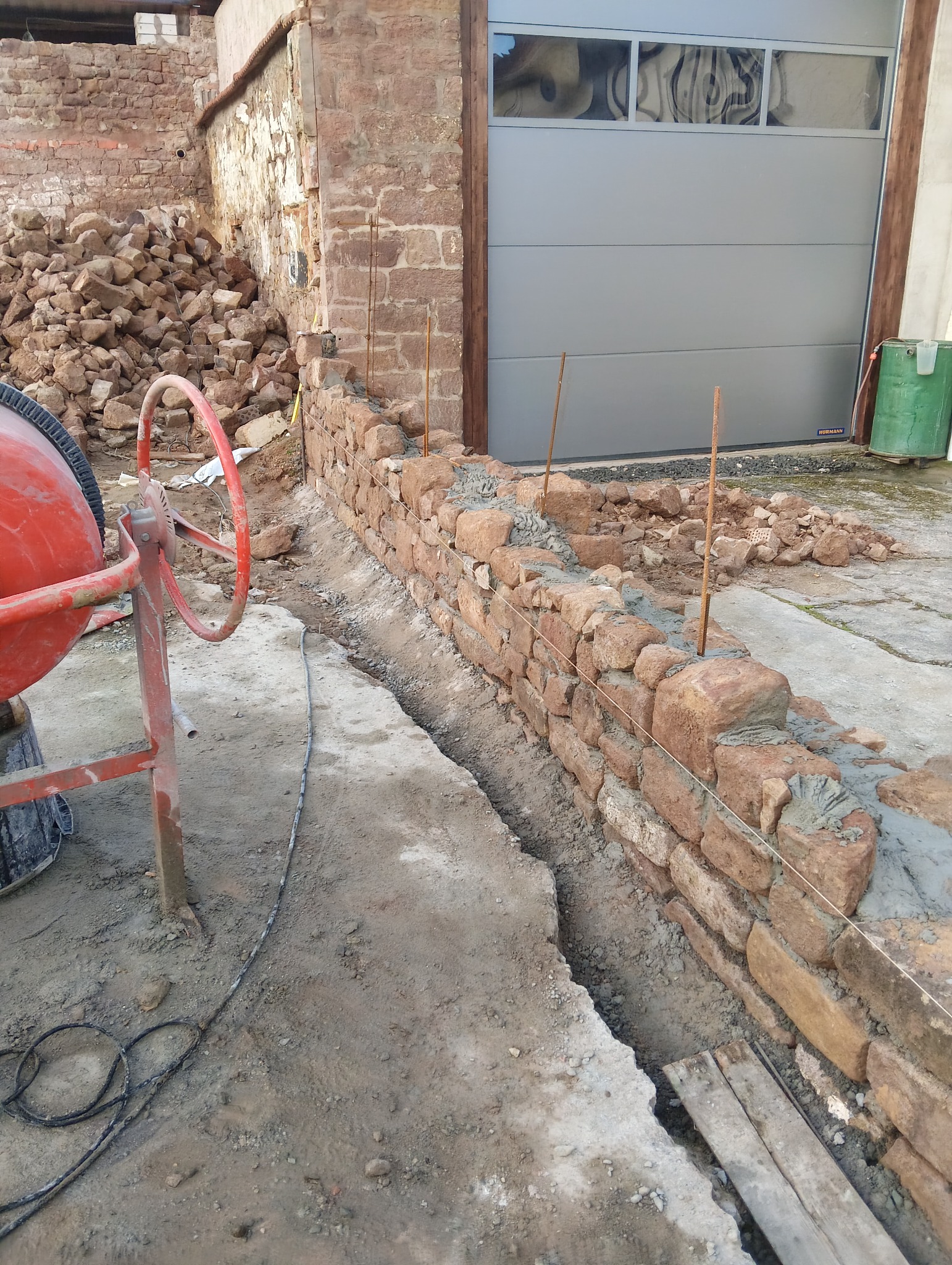 Budowa muru z kamienia polnego. Widoczna zaprawa, pręty zbrojeniowe, betoniarka i stos kamieni. W tle brama garażowa.