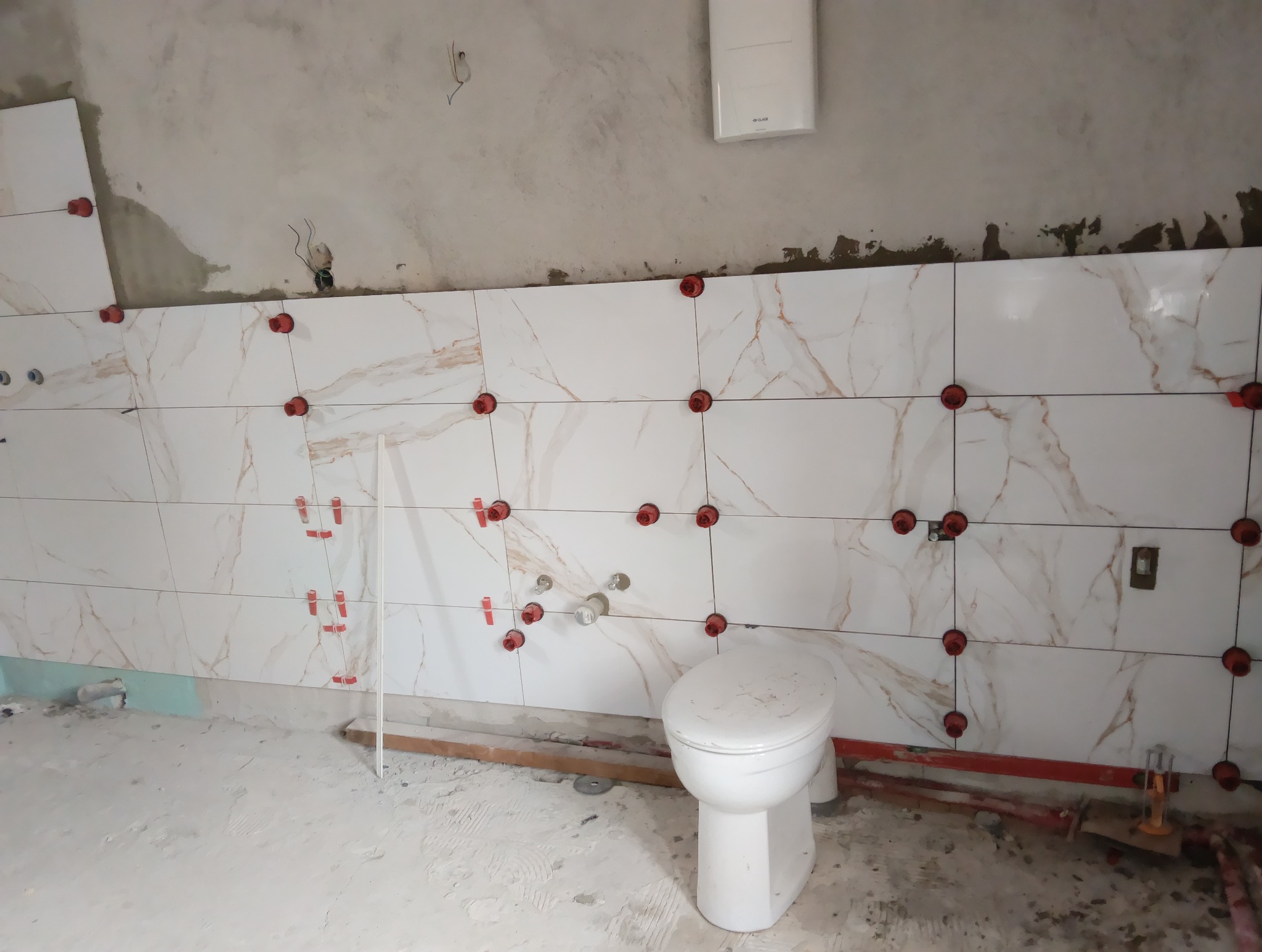 Ściana w trakcie wykańczania z białymi płytkami imitującymi marmur, z czerwonymi elementami poziomującymi, widoczna toaleta i surowy stan podłogi.