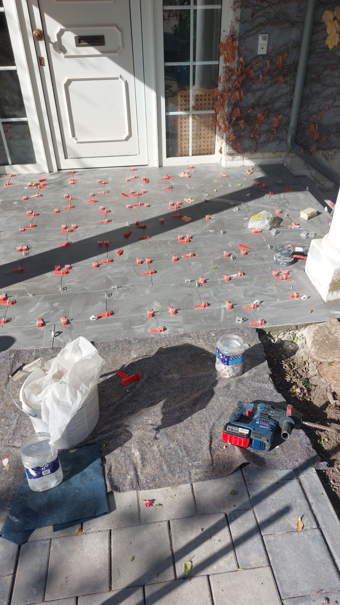 Szare płytki na tarasie z czerwonymi klipsami poziomującymi, białe drzwi wejściowe i bluszcz na ścianie w tle. Widoczne narzędzia i akcesoria do układania płytek.