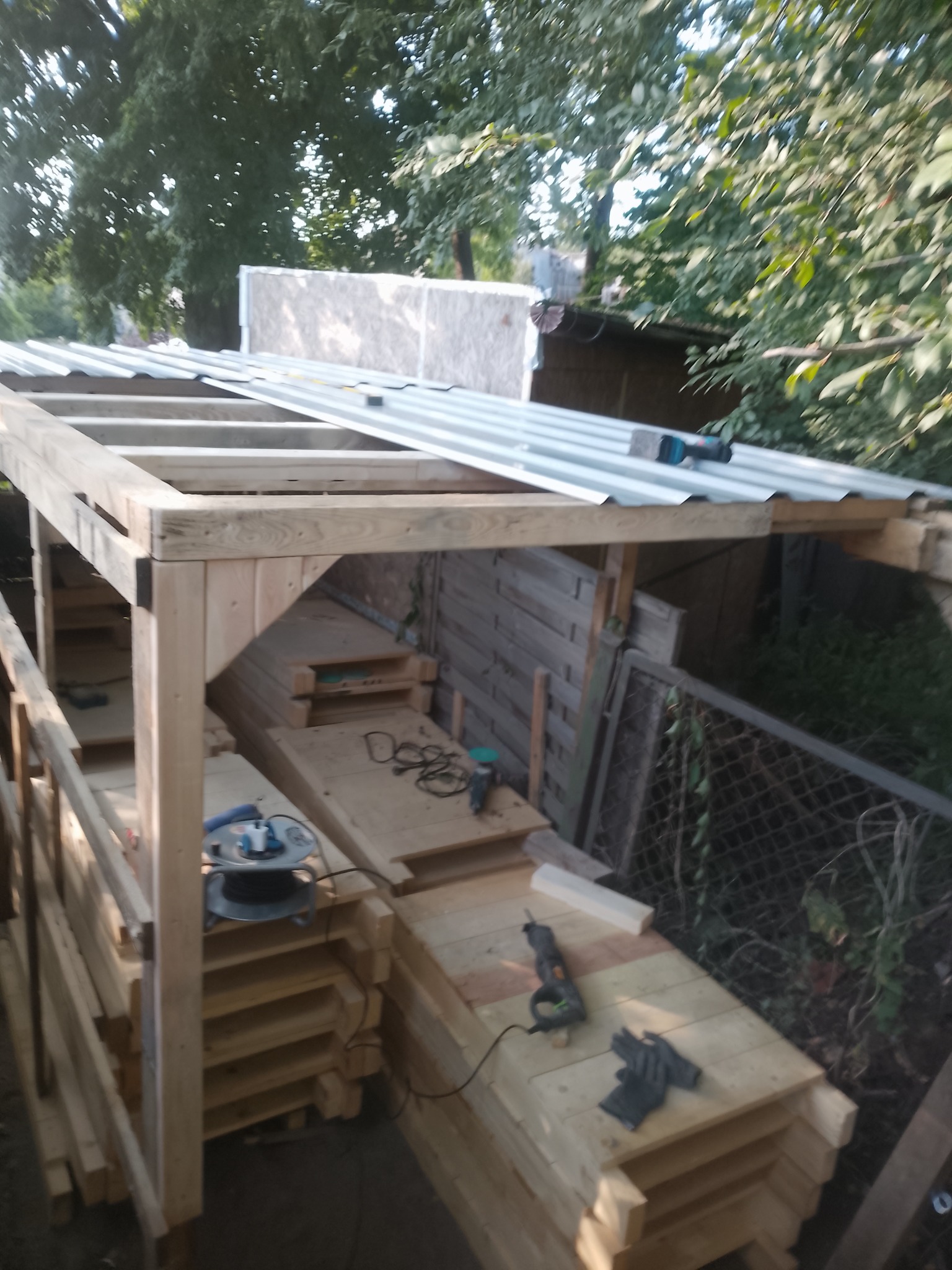 Konstrukcja drewniana z metalowym dachem w ogrodzie, narzędzia i materiały budowlane widoczne. Etap budowy zadaszenia, widok z boku, drzewa w tle.
