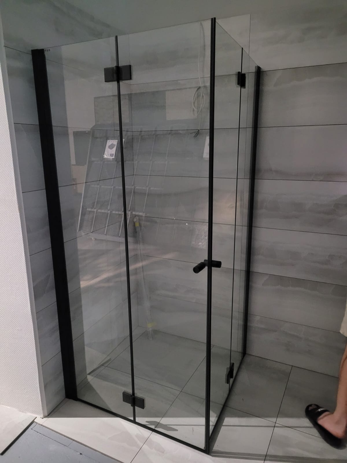 Nowoczesna kabina prysznicowa z czarnymi profilami i przezroczystym szkłem, zamontowana w łazience z szarymi płytkami. Widoczny fragment drabiny w tle.