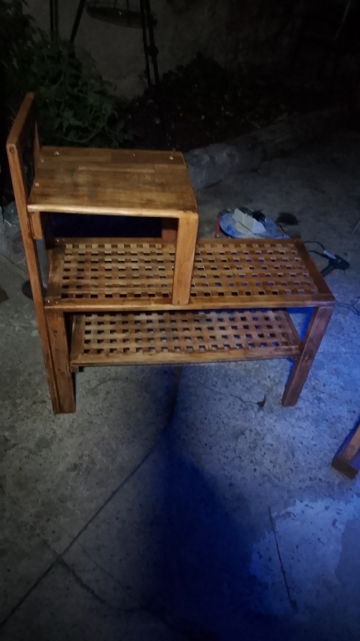 Drewniany, dwupoziomowy regał z ażurowymi półkami, wykonany z ciemnego drewna. W tle widoczne narzędzia i zieleń. Nierównomierne oświetlenie podkreśla fakturę drewna.