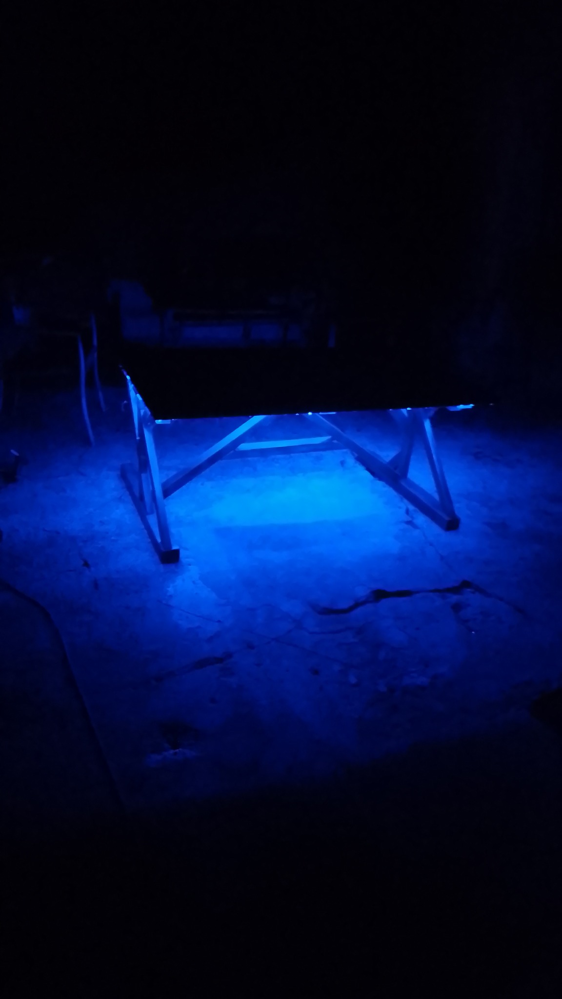 Podświetlony na niebiesko stół z drewnianymi nogami w ciemnym pomieszczeniu. Nierówna podłoga podkreślona przez intensywne, niebieskie światło.