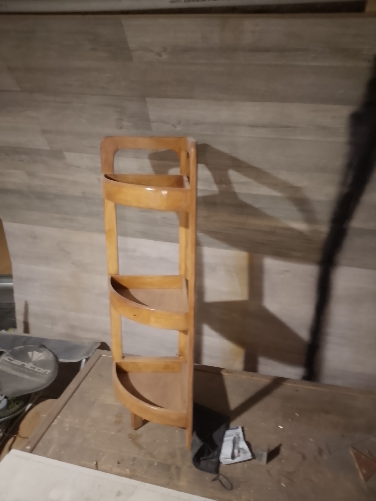 Drewniany, półokrągły regał z trzema półkami, stojący na podłodze z widocznym tłem w postaci paneli ściennych. Ciemne oświetlenie, rzucające cień na ścianę.