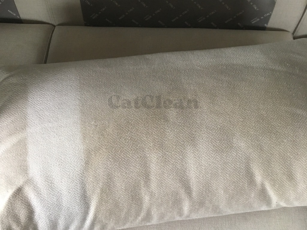 Jasna poduszka z delikatnym wzorem jodełki i widocznym napisem 'CatClean', leżąca na jasnej sofie. Widoczna różnica w czystości poduszki.