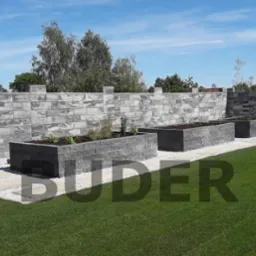 Nowoczesny ogr&oacute;d z szarego kamienia: podwyższone rabaty i mur oporowy, ścieżka z jasnych płyt, trawnik i błękitne niebo.