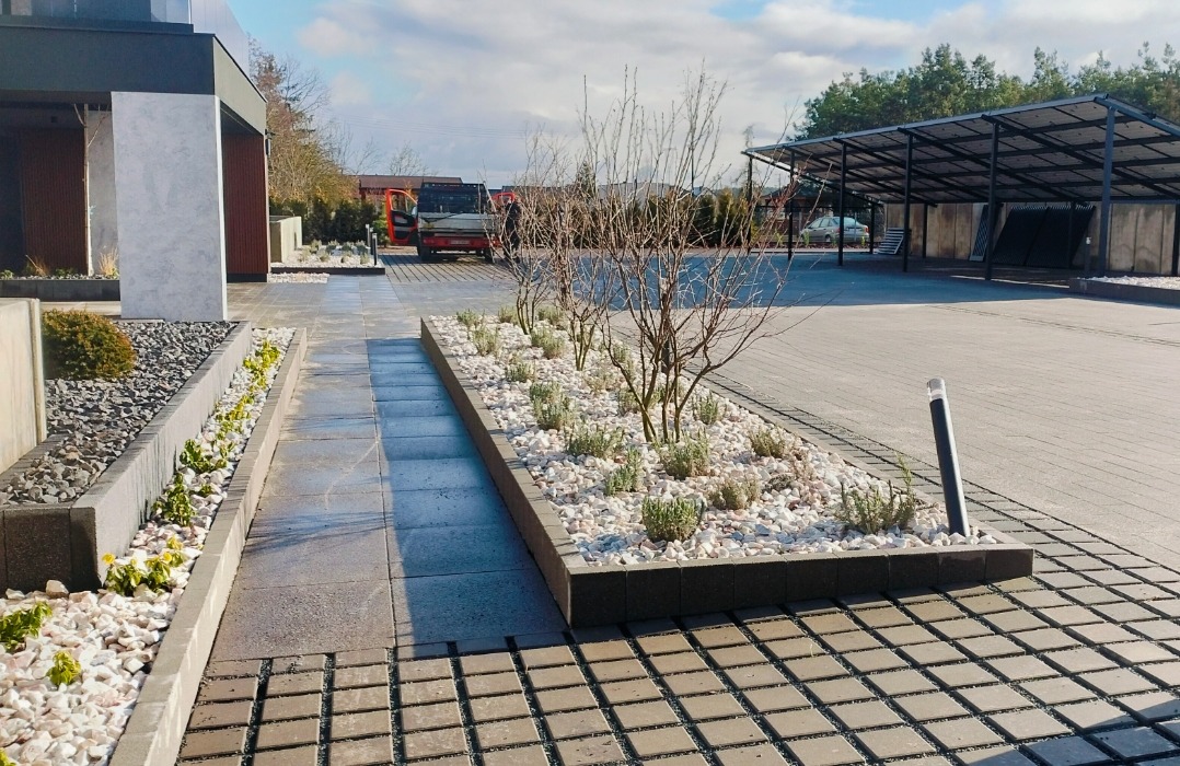 Ułożona kostka brukowa w Santoku z rabatami obsypanymi białym kamieniem i niską zielenią, na tle wiaty parkingowej i zaparkowanego samochodu dostawczego.