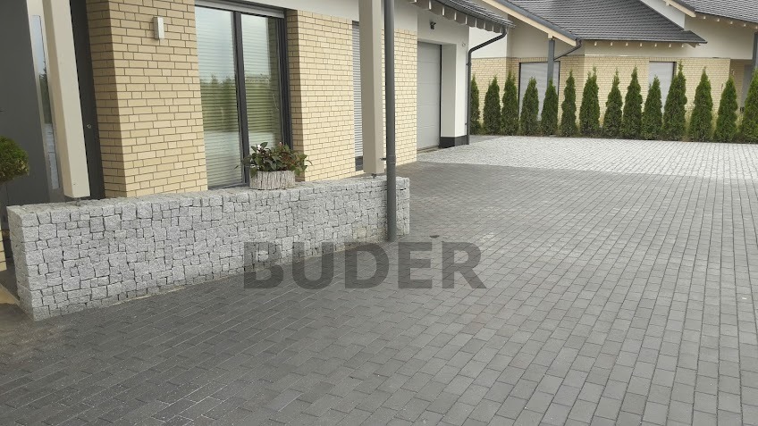 Szara kostka brukowa ułożona na podjeździe przed domem z cegły, z niskim murem z kamieni i rzędem tuj w tle.