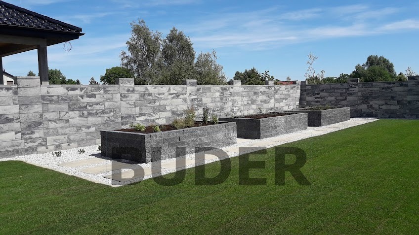 Nowoczesny ogród z szarego kamienia: podwyższone rabaty i mur oporowy, ścieżka z jasnych płyt, trawnik i błękitne niebo.