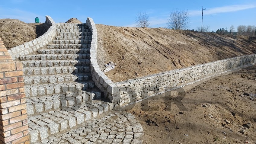 Granitowe schody prowadzące na wzgórze, wykończone murem oporowym z kamienia, widoczne z poziomu ziemi w słoneczny dzień.