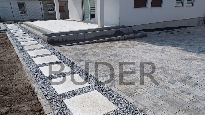 Ułożona ścieżka z kwadratowych płyt i szarego żwiru prowadząca do tarasu z kostki brukowej przy białym domu.