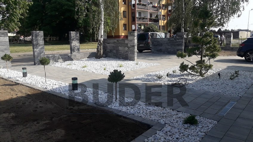 Nowoczesny podjazd z szarej kostki brukowej, ozdobiony białymi kamieniami i miniaturowymi drzewkami, otoczony kamiennymi słupkami ogrodzenia.