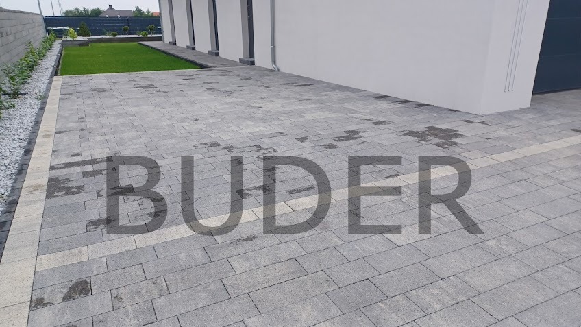 Ułożona szara kostka brukowa z jasnym pasem oddzielającym od trawnika i domu, widoczne ślady wilgoci na powierzchni.