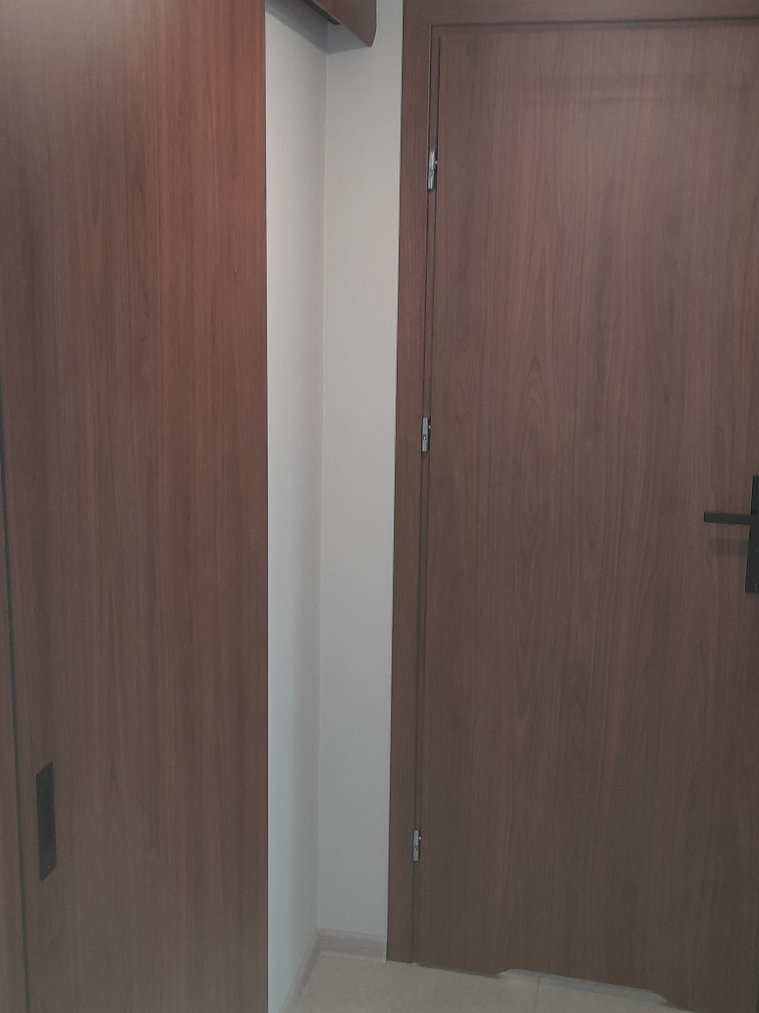 Zamknięte, brązowe drzwi wewnętrzne z czarną klamką, widoczne obok fragmentu szafy wnękowej o podobnym wykończeniu, na tle białej ściany i jasnej podłogi.