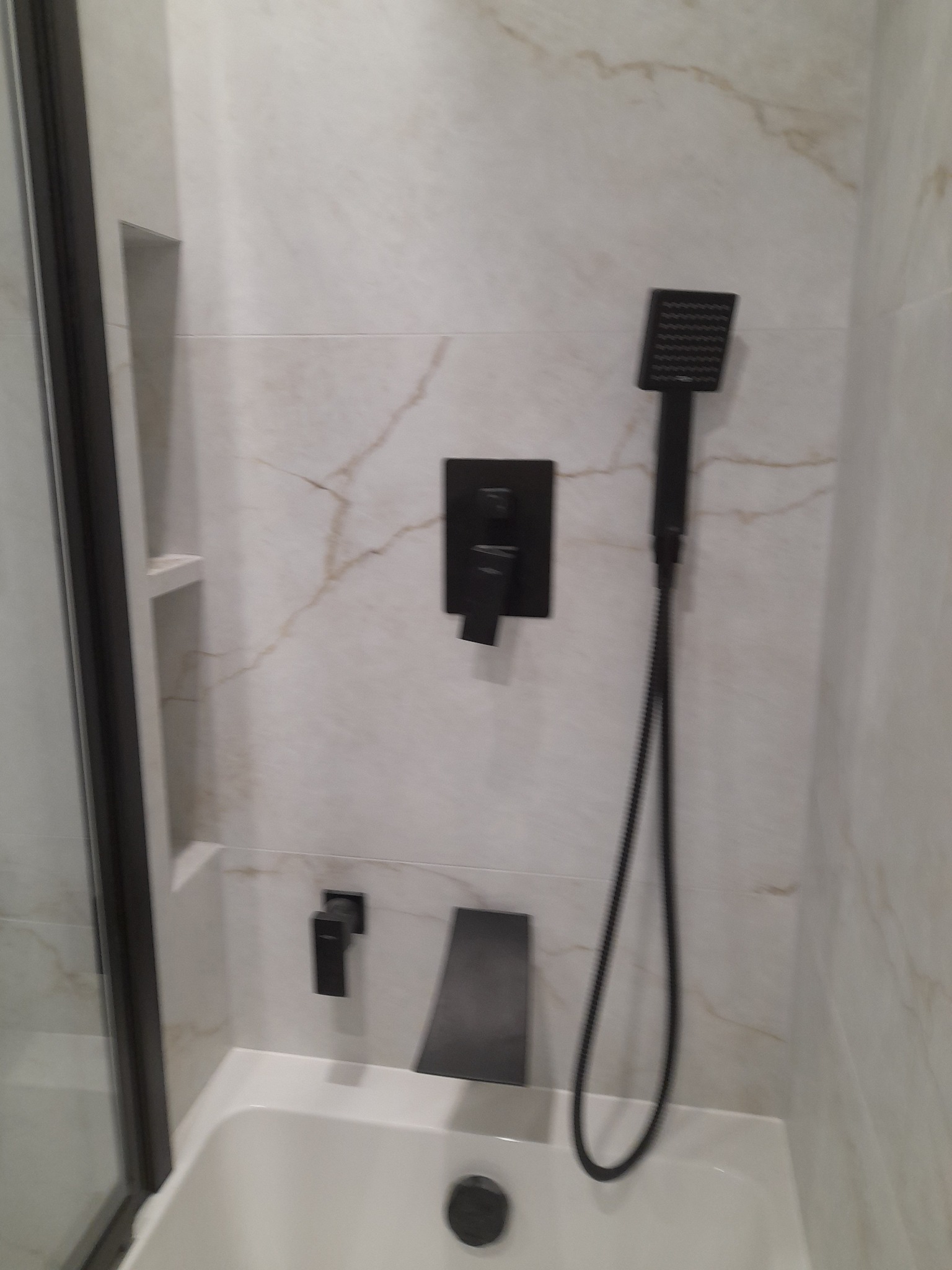 Nowoczesna armatura w wykończonej łazience w Lublinie. Bateria wannowa i deszczownica w kolorze czarnym, na tle płytek imitujących marmur.